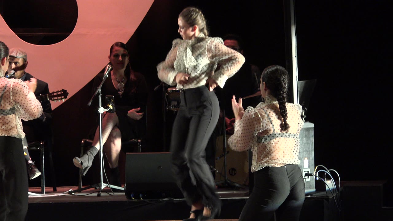 At 0:01 in Cabo San Lucas, Mexico, the central dancer turns with hands near her waist as a foreground dancer claps and seated musicians remain in the background.
