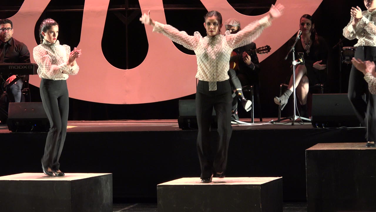 A woman in dark pants stands on a square platform during a tap dance performance in Cabo San Lucas, Mexico, with seated musicians and pink stage decor behind her.