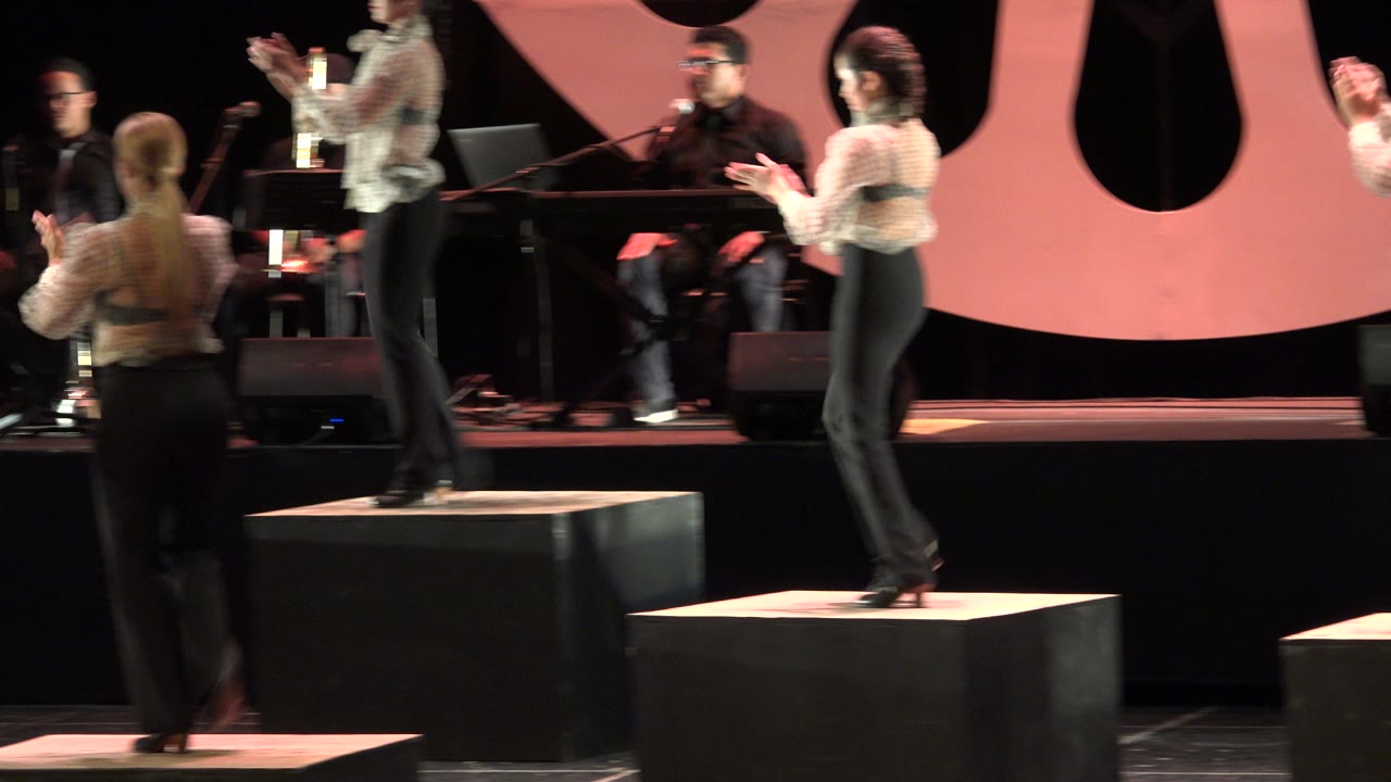 Performers clapping on black stage boxes with musicians behind them.