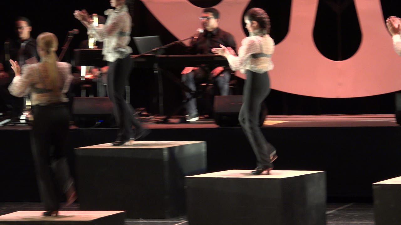 In Cabo San Lucas, Mexico, multiple performers clap from separate black platforms as a band sits behind them beneath warm lighting and a pink stage backdrop.