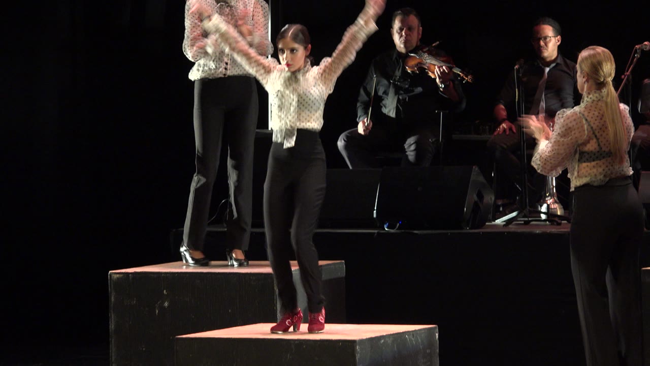 Young girl in a ruffled light blouse and black pants performs on a dark stage in Cabo San Lucas, Mexico, with seated musicians and another performer behind her.