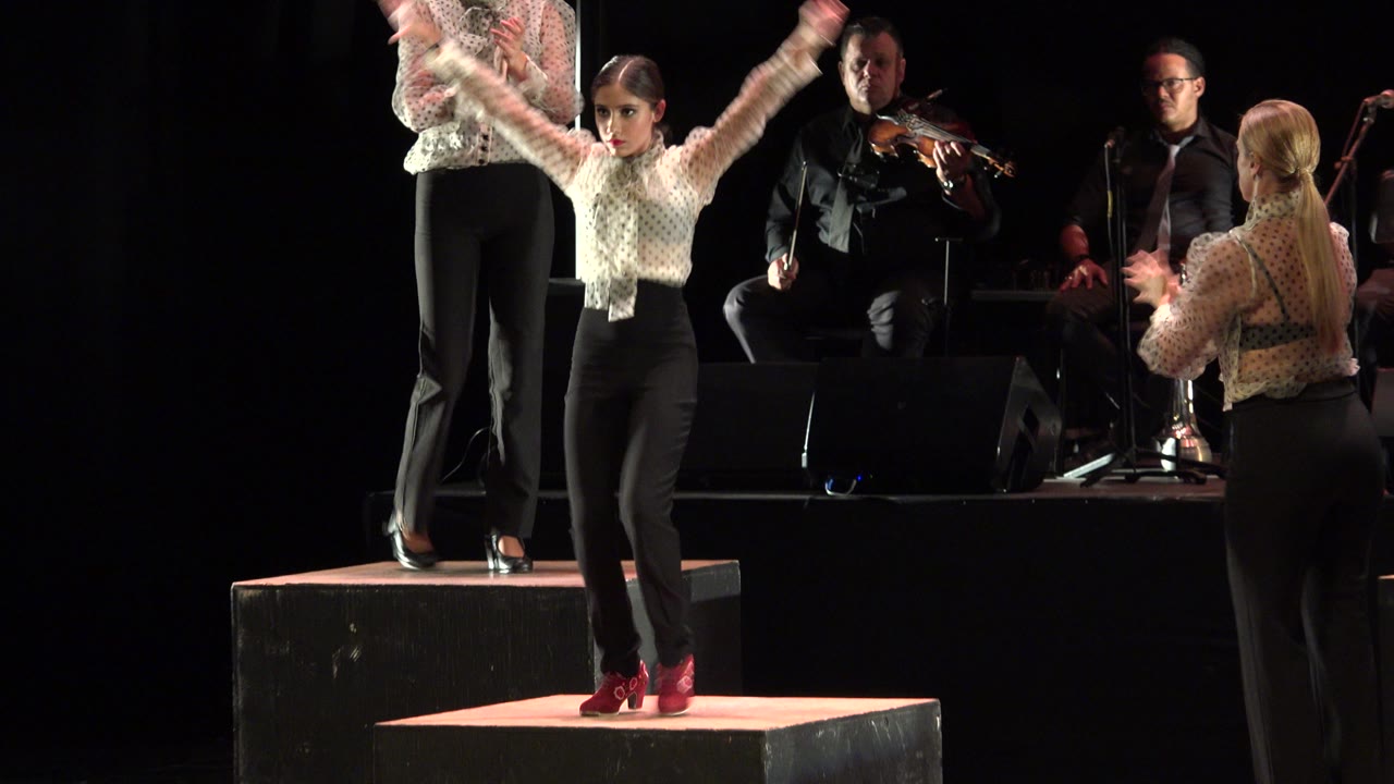 Child dancer raises both arms while facing forward on the Cabo San Lucas stage, with a violinist and another seated musician visible in the background.