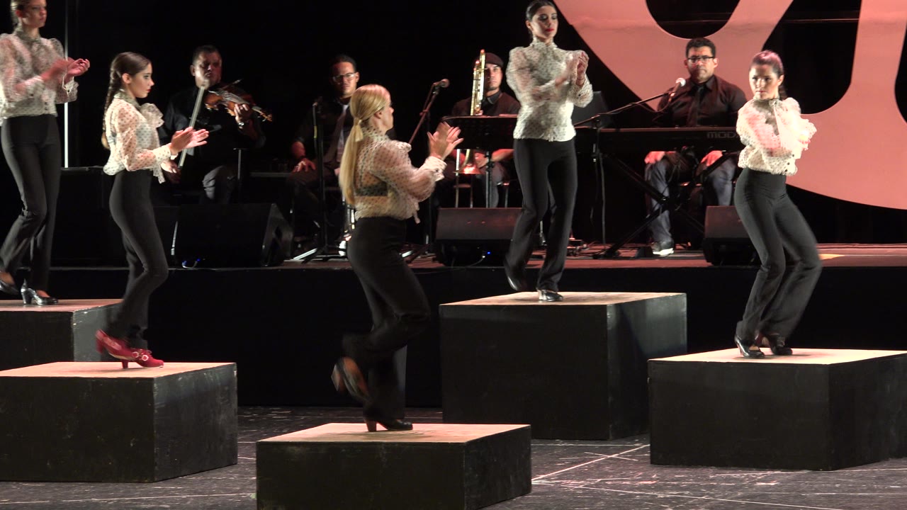At 0:00 in Cabo San Lucas, women in white blouses and black trousers clap and step on black stage risers while musicians sit behind them.