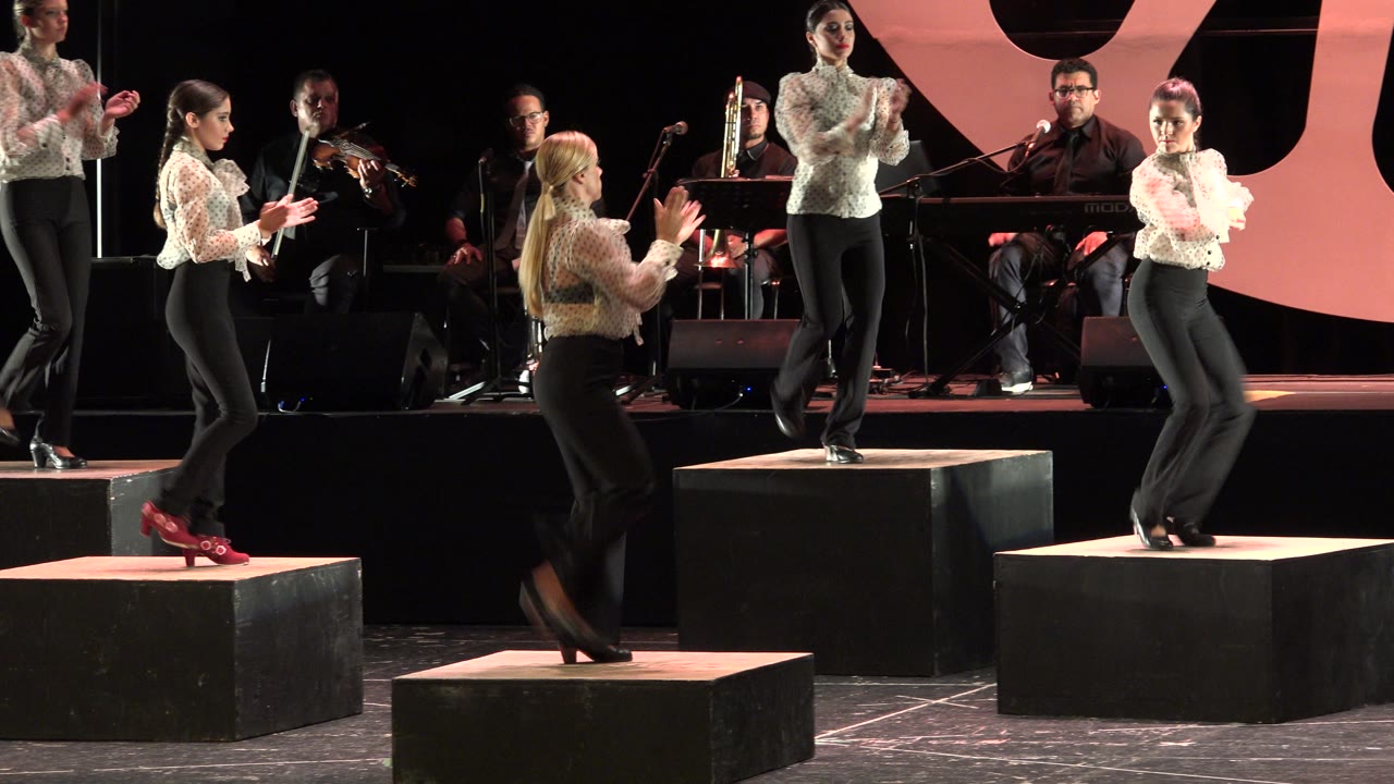 At 0:01 in Cabo San Lucas, the dancers continue clapping in formation on separate black platforms, with a violinist and seated musicians in the rear stage area.