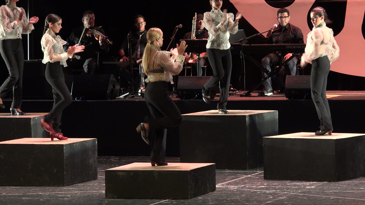 Dancers clap and step on black stage risers with musicians behind them.