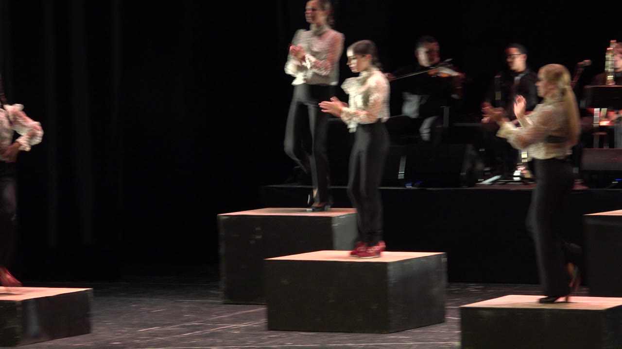 Wide stage view in Cabo San Lucas showing women in white blouses and black pants dancing on raised black platforms, with seated musicians partly visible in the dark background.