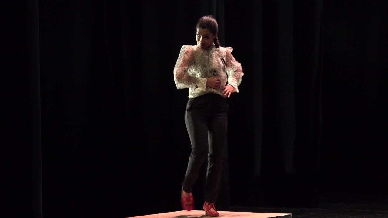 At 0:01, the dancer remains centered on the raised box, facing forward with one hand near the waist and the dark curtain behind.
