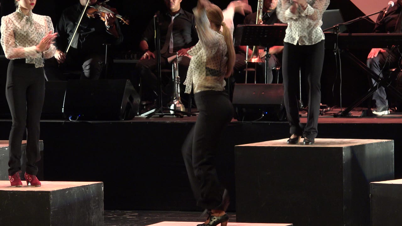 Stage performers clap beside a raised platform with musicians behind them.