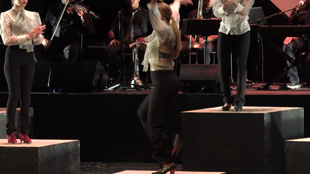 A male dancer in dark pants crosses the Cabo San Lucas stage between clapping performers, with seated musicians and music stands behind him.