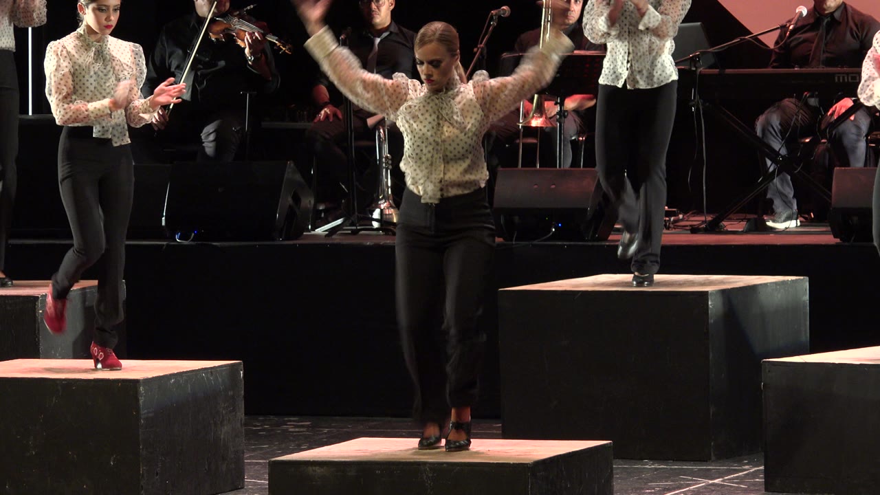 In Cabo San Lucas, the central dancer faces forward with arms blurred in motion, standing between raised black stage blocks as musicians remain visible behind.