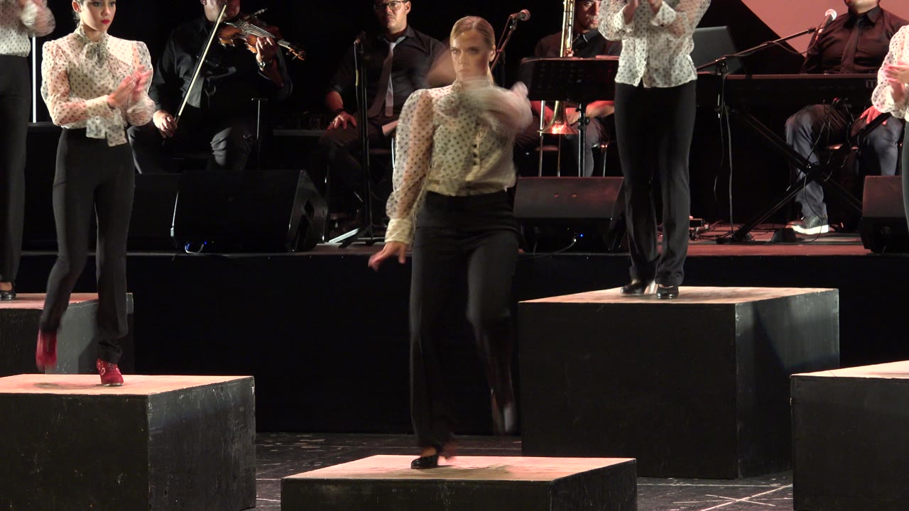 Young woman tap dancing on black stage blocks with performers behind her.