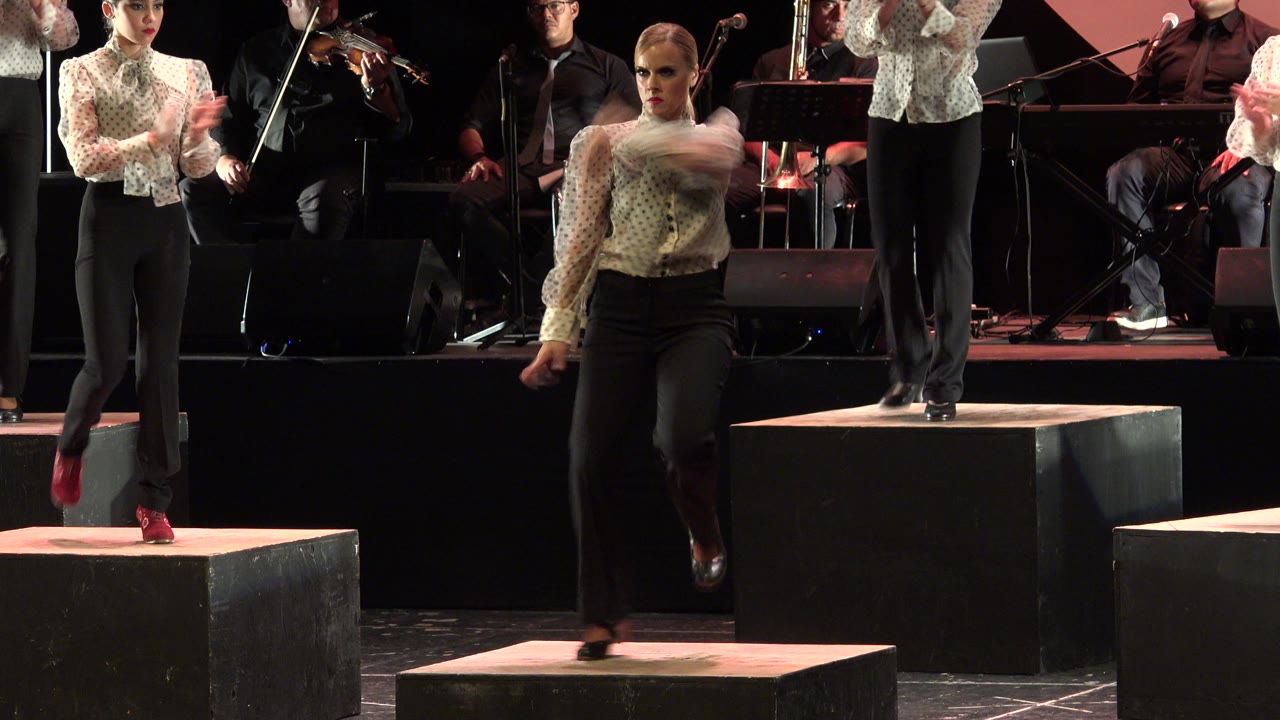 At 0:01 in Cabo San Lucas, the dancer steps forward with one leg blurred by motion while another performer claps on a raised stage block behind her.