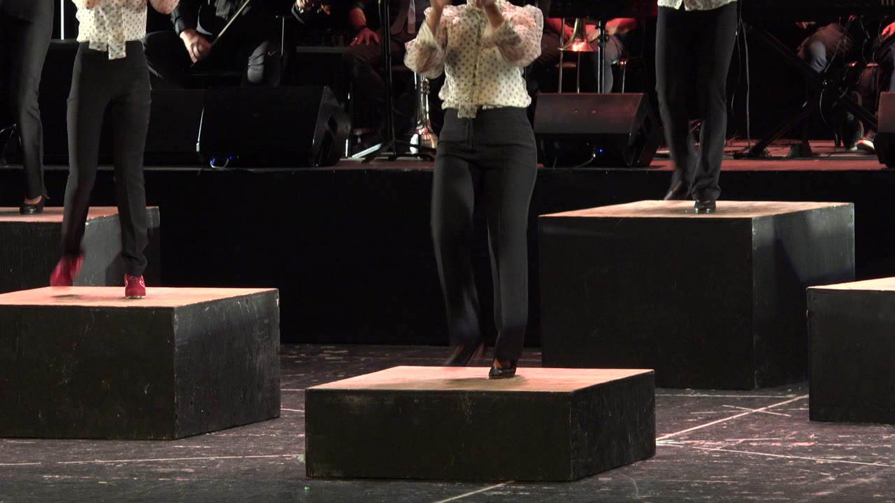 In Cabo San Lucas, Mexico, cropped performers stand and step on black stage platforms while musicians and stage equipment sit in the dark background.