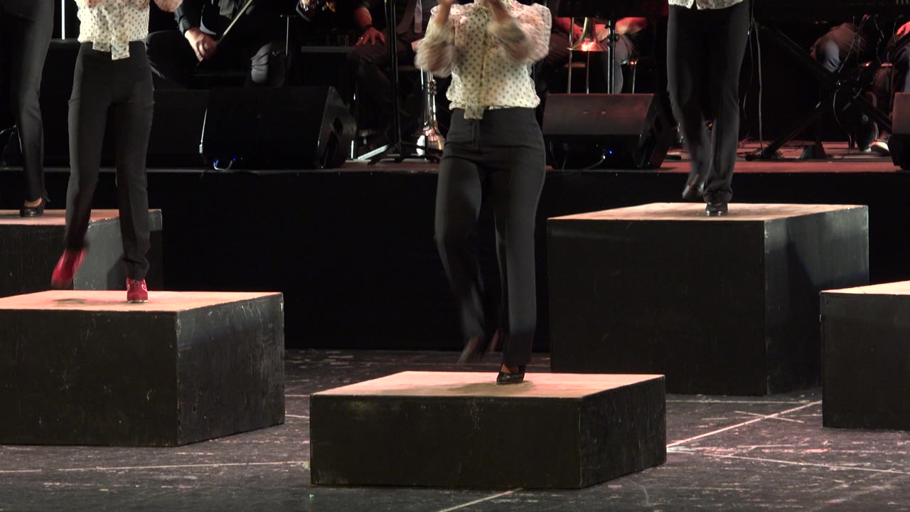 A cropped dancer steps onto the front black platform in Cabo San Lucas while other performers and stage musicians remain partly visible behind.
