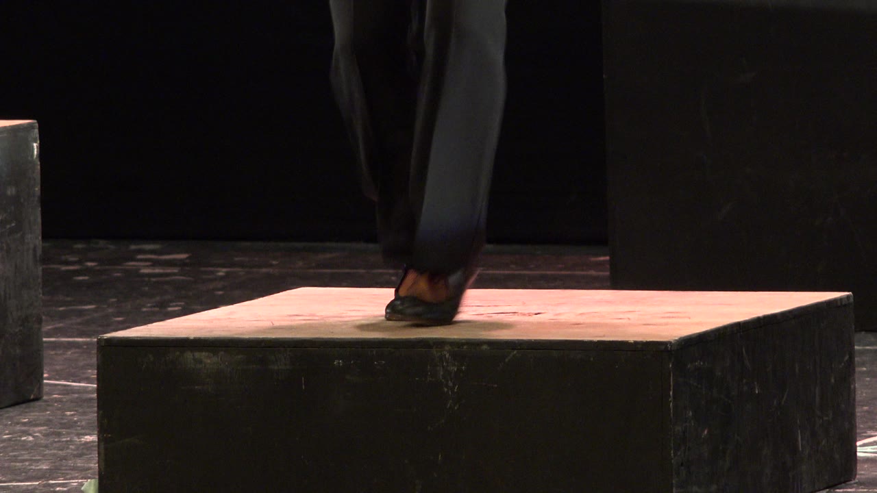 One second in, the Cabo San Lucas performance shows the dancer's trouser legs crossing tightly above the wooden board, with black stage blocks behind.