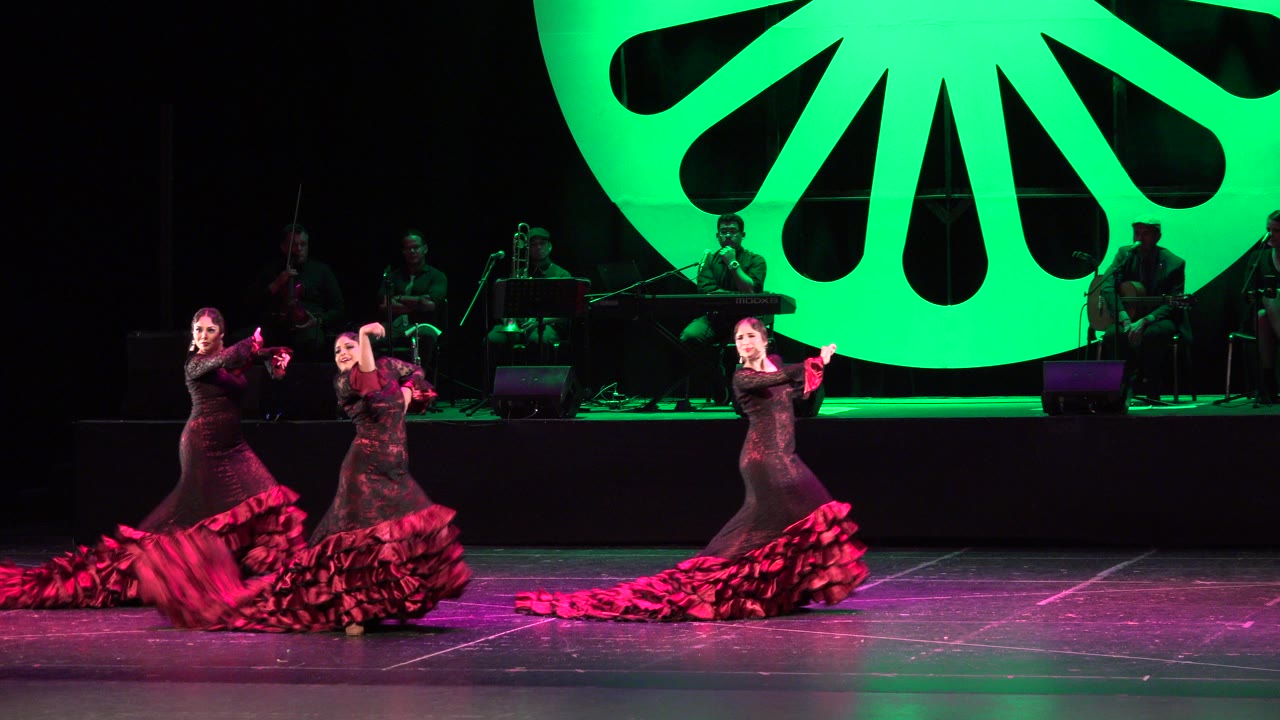 Three women dancing on a stage with green backdrop and musicians behind them.
