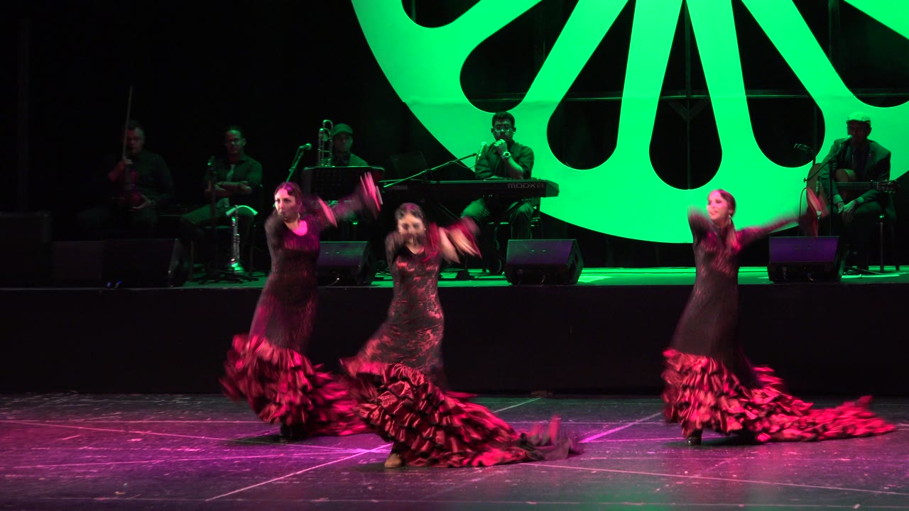 Three women dance on a stage with musicians and a green backdrop behind them.