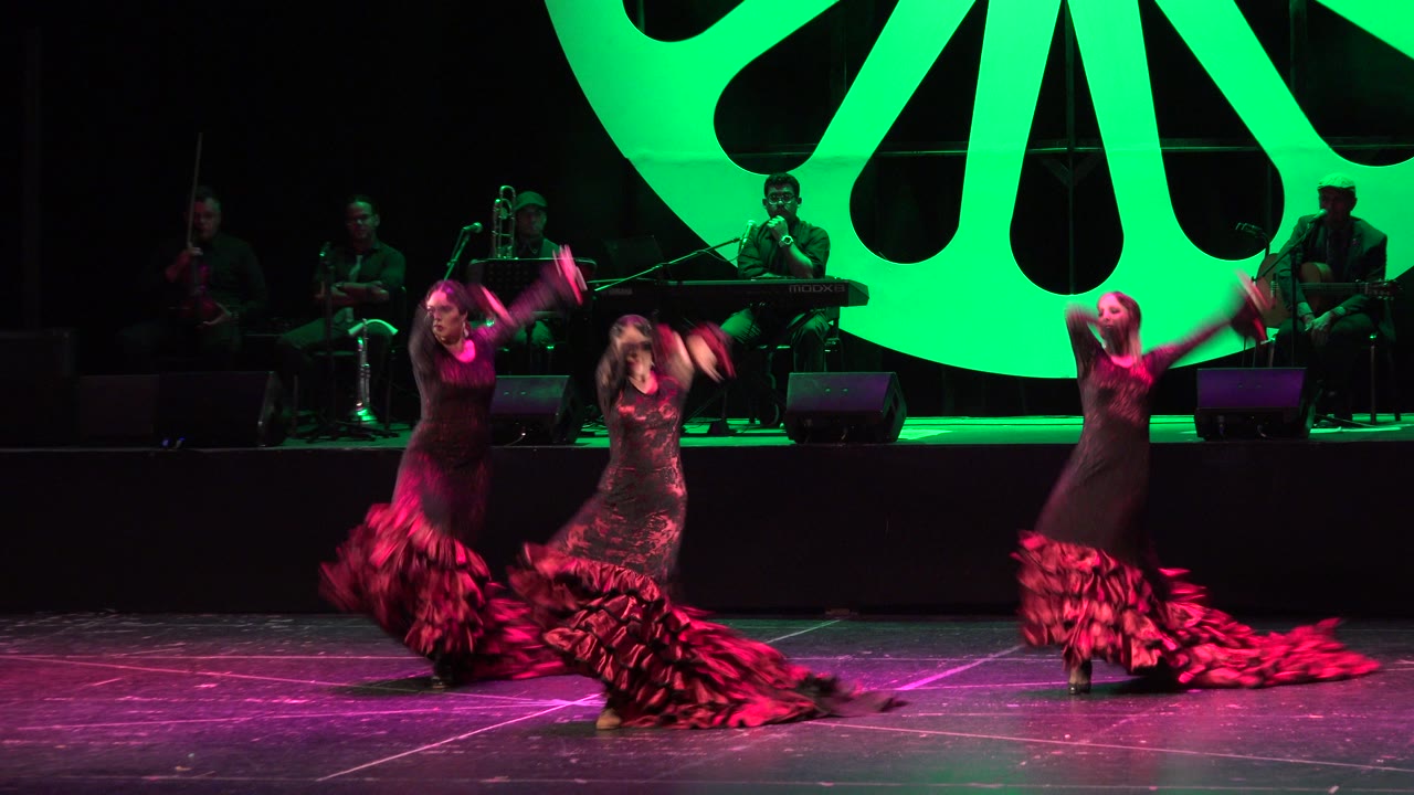 Three dancers in matching dark dresses move through a choreographed moment on a Cabo San Lucas stage as red skirt ruffles spread across the floor.