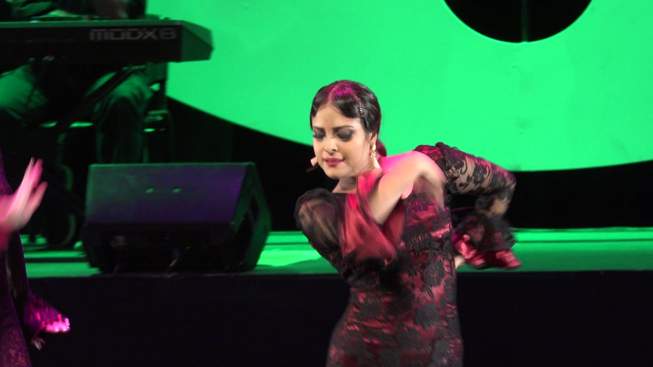 The Cabo San Lucas stage dancer holds a low forward pose with one arm lifted, backed by green light, dark stage flooring, and visible sound equipment.