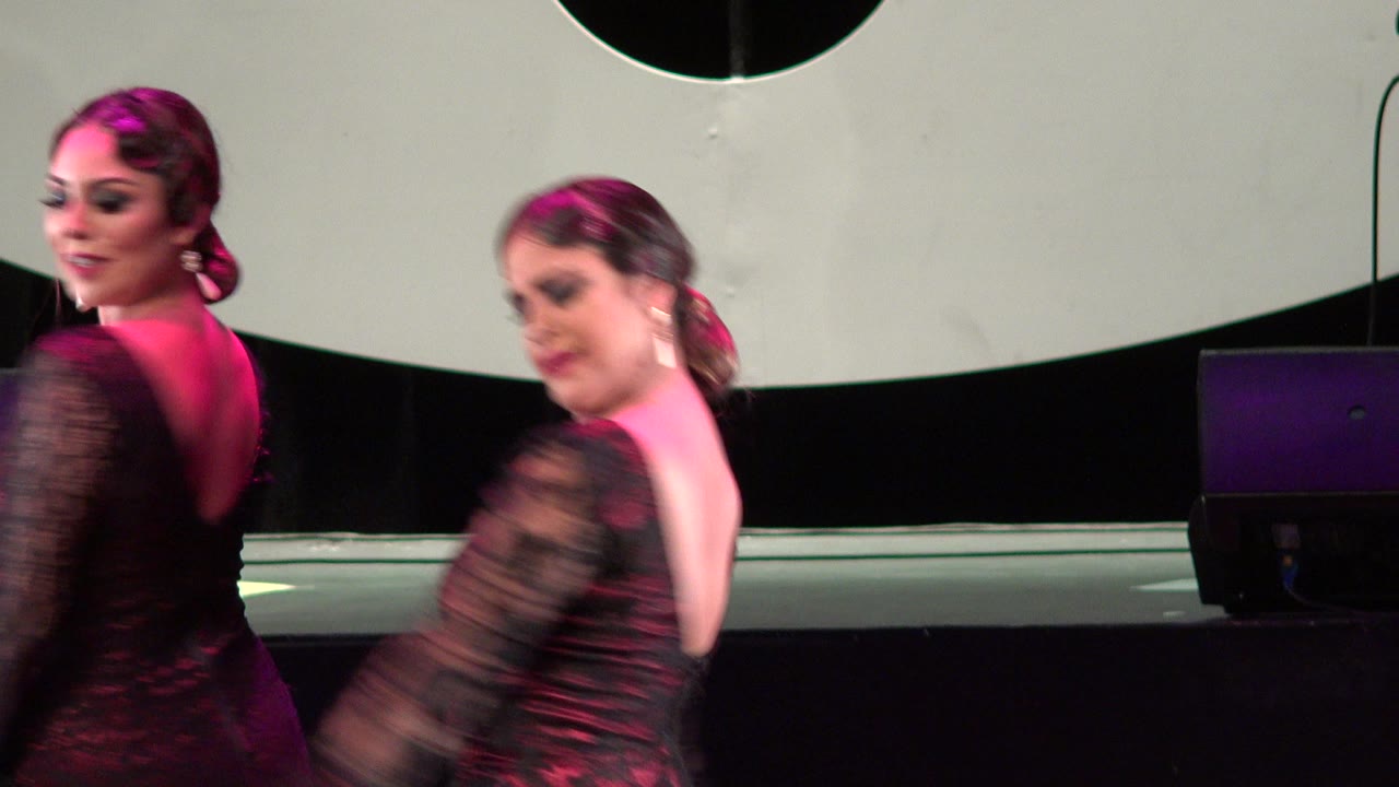 Two adult women in red and black flamenco-style dresses dance on a stage in Cabo San Lucas, Mexico, with pink light on their hair and shoulders.