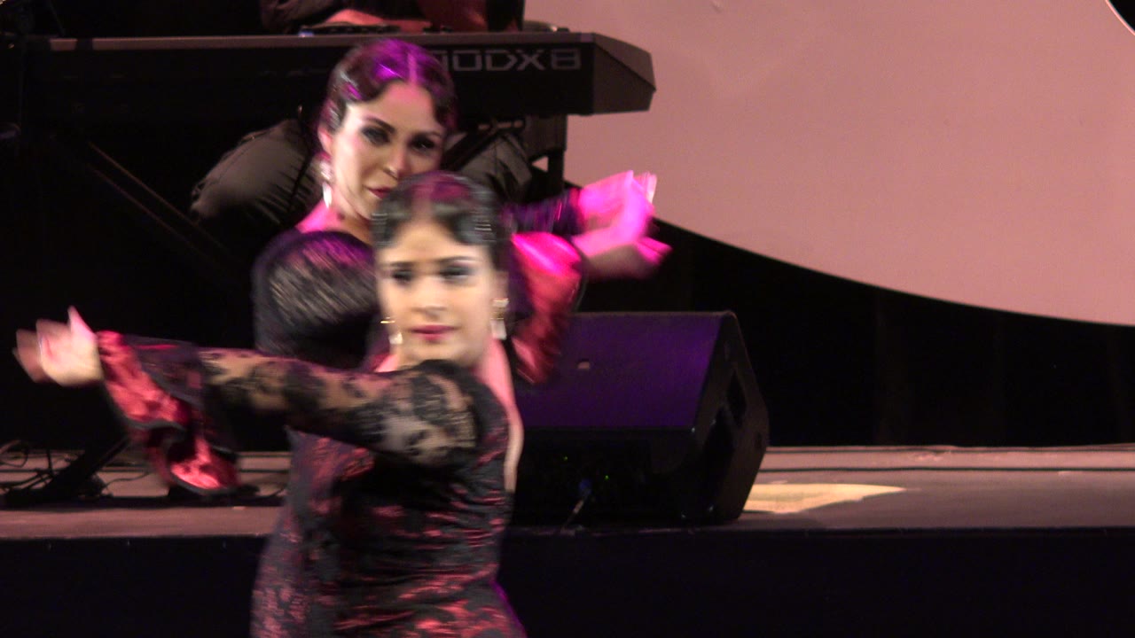 Two performers continue the Cabo San Lucas stage dance, with one face turned toward camera, blurred arms in motion, and a keyboard visible at the rear.