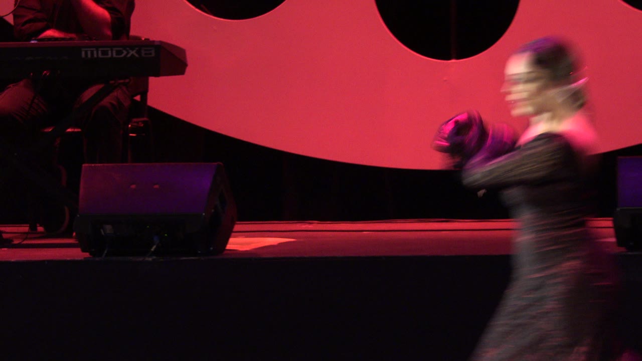 A dancer appears motion-blurred near center frame on the red-lit stage in Cabo San Lucas, Mexico, while a shadowed keyboard player sits at the back left.