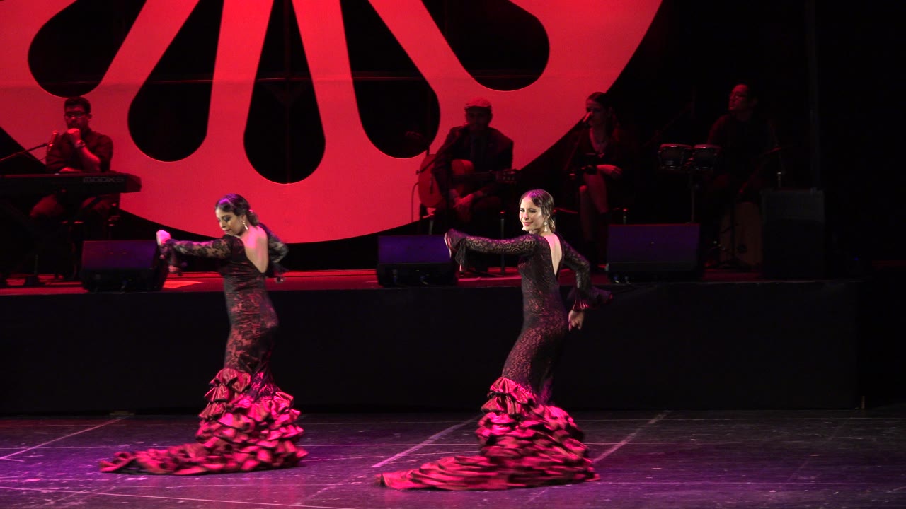 Two women dance on a red-lit stage with musicians behind them.