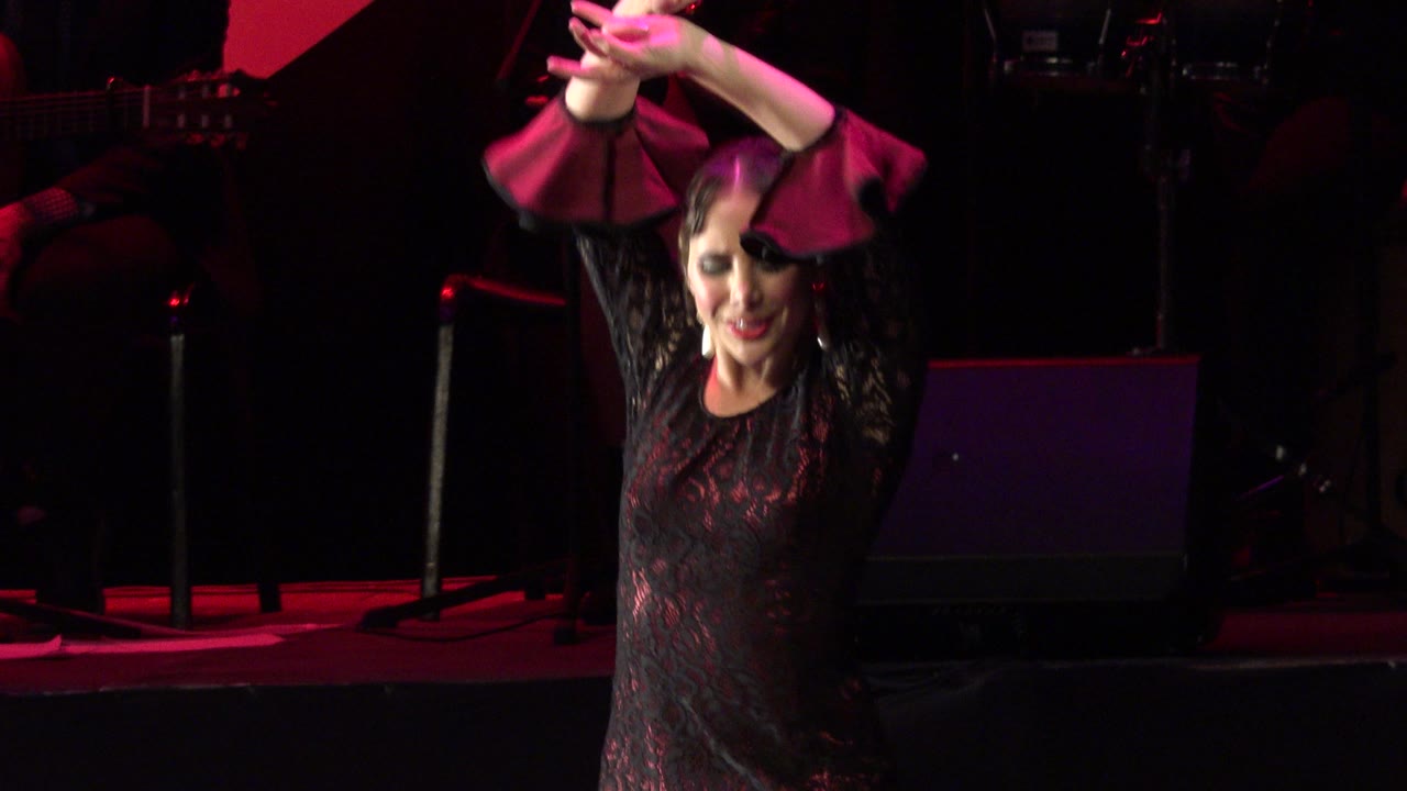 The dancer keeps both hands above her head as pink-red light catches the sleeves of her dark dress on the Cabo San Lucas stage.