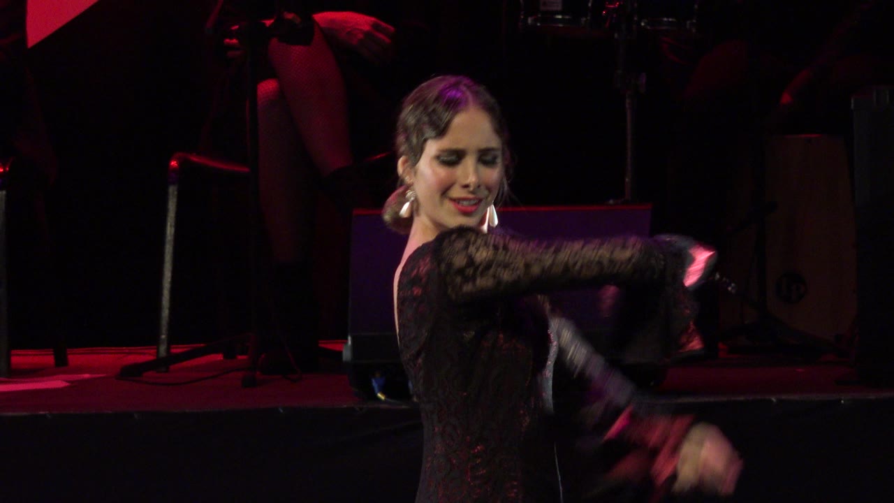 Female dancer in a black lace dress performs on a dark stage in Cabo San Lucas, Mexico, with red floor light and percussion equipment behind her.