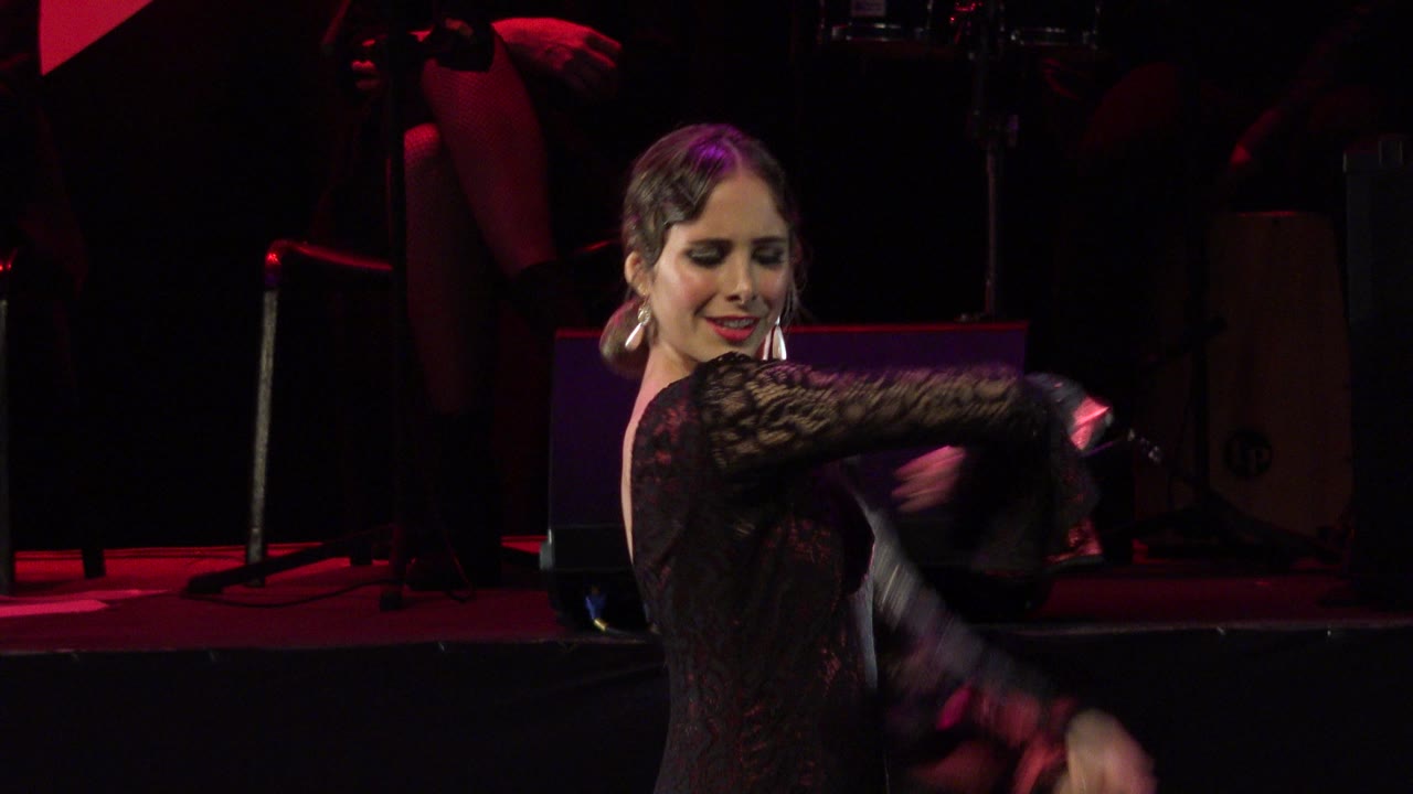 Female performer in Cabo San Lucas, Mexico, moves her arms in front of a dark stage backdrop, with red lighting and background instruments visible.