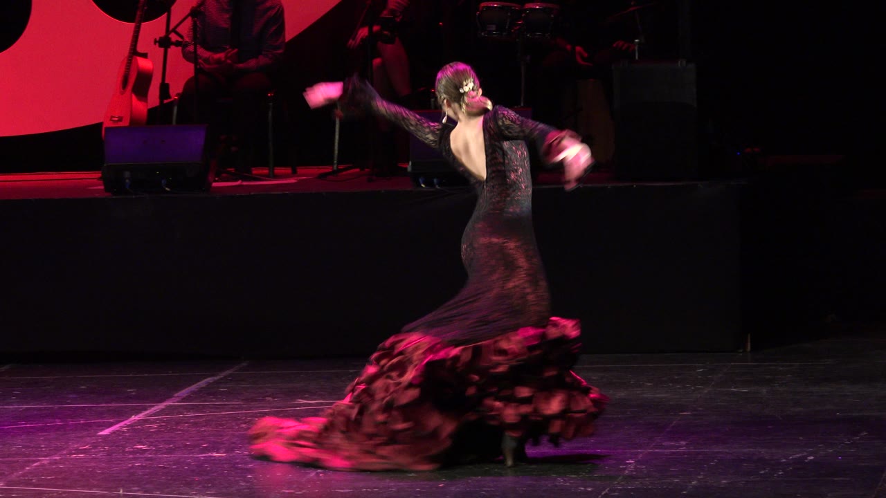 The dancer continues turning in Cabo San Lucas, Mexico, with her arms extended and the red ruffled hem blurred by motion across the black stage floor.