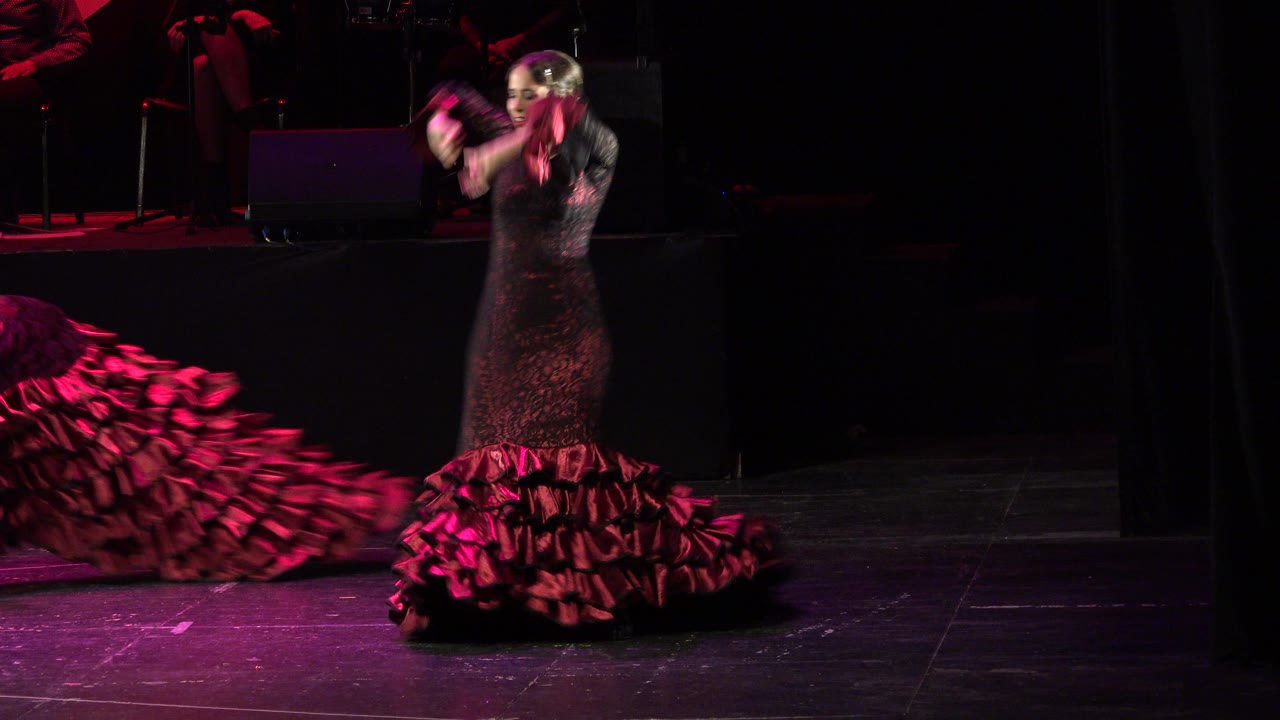 In Cabo San Lucas, Mexico, the woman continues dancing with lifted hands, her red ruffled dress gathered around her feet on the dark stage.