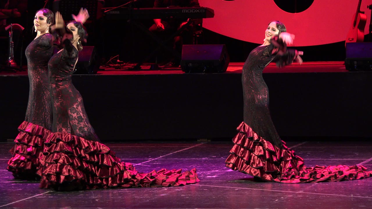 Two adult flamenco dancers in Cabo San Lucas face the audience area as one extends her arm and the other turns, with red lighting and a dark equipment riser behind them.
