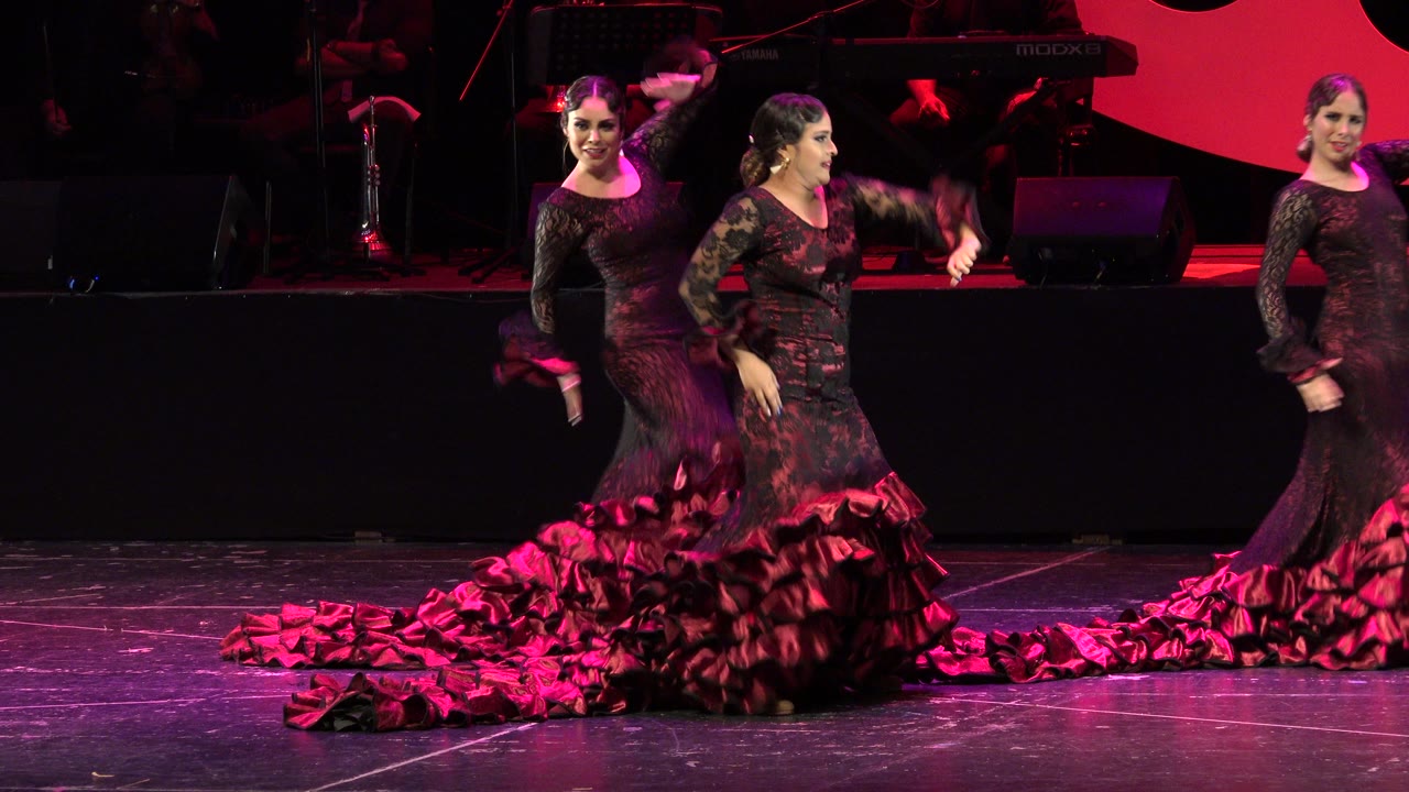 Three women in black lace dresses and ruffled red skirts perform on an indoor stage in Cabo San Lucas, Mexico, with microphones and stage gear behind them.