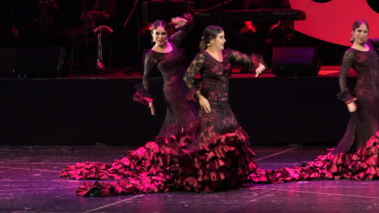 The dancers remain in formation on the Cabo San Lucas indoor stage, with one performer facing forward and another turning beside a long red ruffled train.