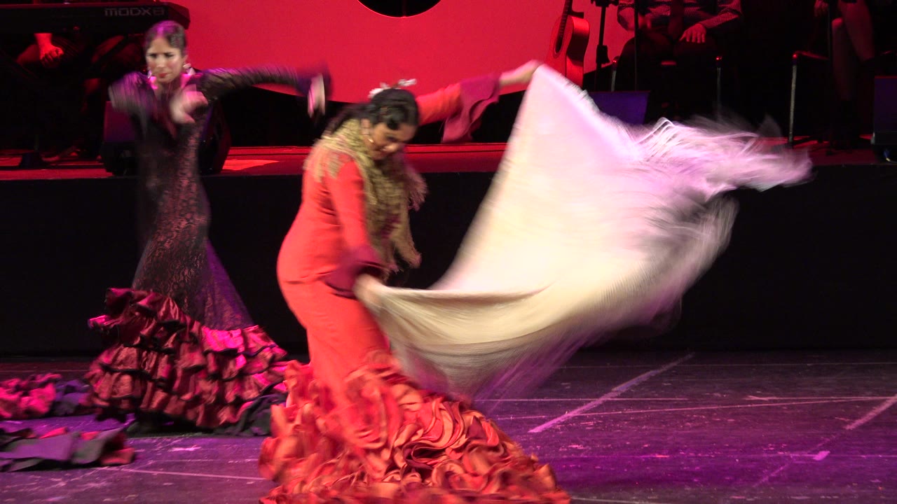 Two women in ruffled dresses dance as a white shawl swings across the stage.
