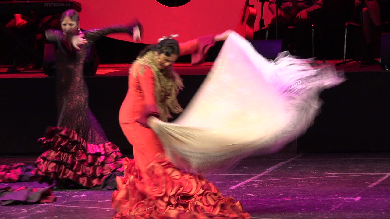 A woman in a red ruffled dress bends forward with a white fringed shawl as a second dancer in black and red moves behind her on the Cabo San Lucas stage.