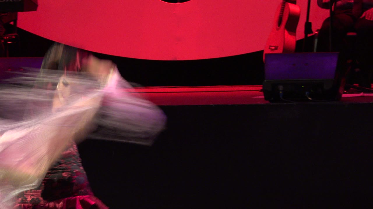 At 0:01 in Cabo San Lucas, Mexico, the dancer is partly visible behind a blurred shawl or skirt sweep, with a red backdrop and dark stage equipment behind her.