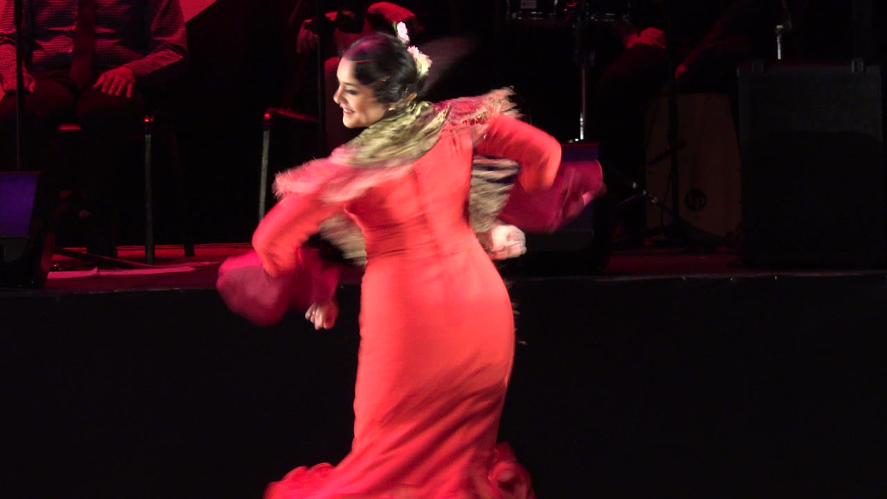 A woman in a red ruffled dress turns on a live stage in Cabo San Lucas, Mexico, with red lighting and dark stage equipment behind her.