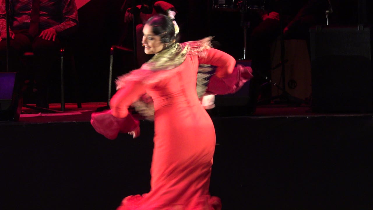 The dancer remains partly turned away during the Cabo San Lucas stage performance, with her red dress lit against a black background and instruments behind her.