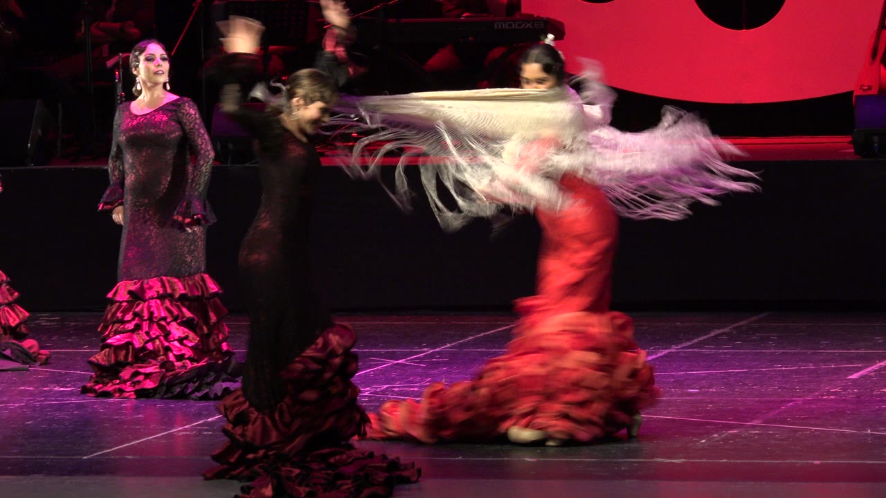 A red-costumed flamenco dancer in Cabo San Lucas moves across the stage with a white shawl blurred by motion, while two black-dressed dancers remain in view.