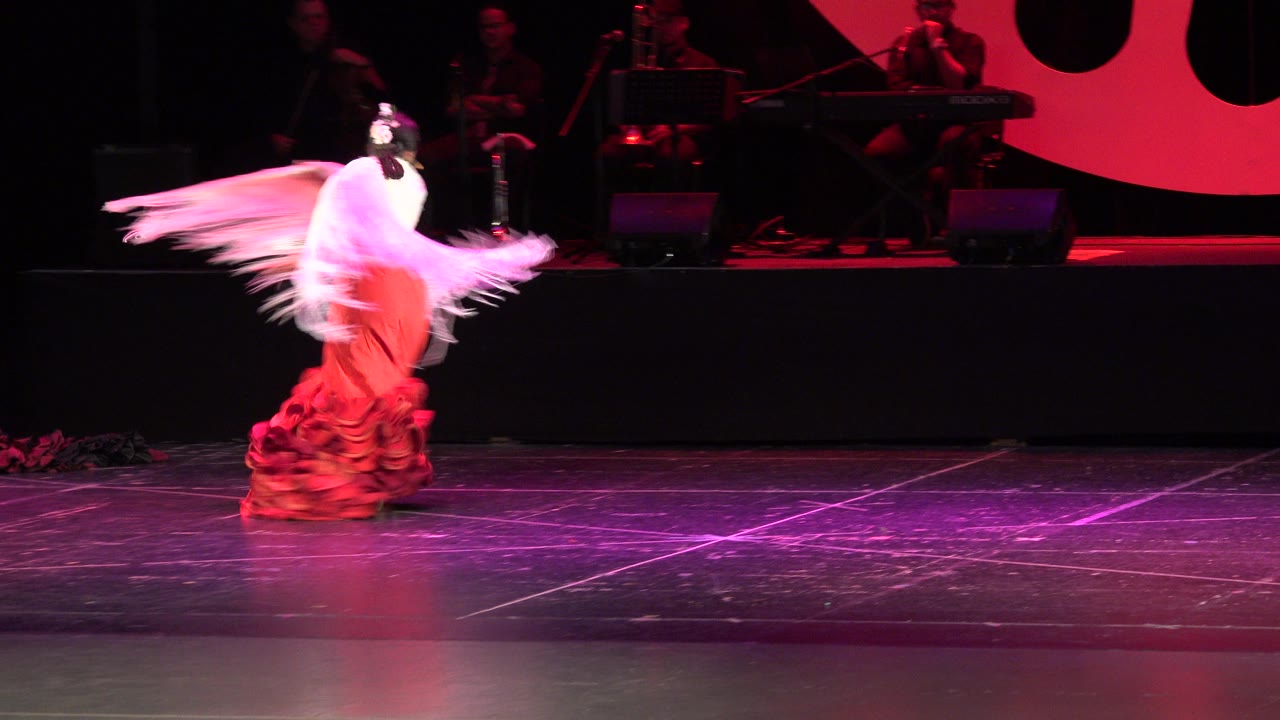 Solo dancer in red dress swings a pale shawl on a dark stage.