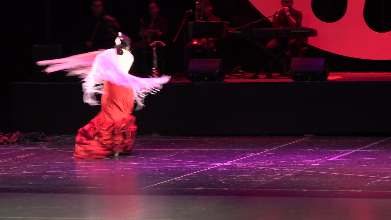 The solo dancer turns near center stage in Cabo San Lucas, Mexico, with the pale shawl stretched behind her and shadowed musicians on the rear platform.