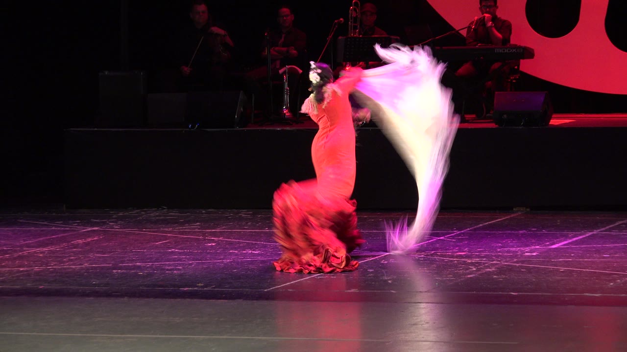 In Cabo San Lucas, Mexico, the red-dressed dancer faces partly away while lifting the white scarf into a tall curve beside the dark stage backdrop.