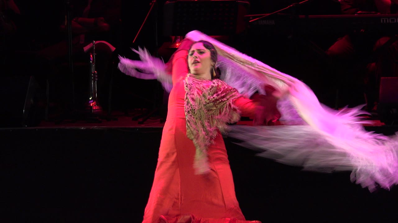 In Cabo San Lucas, Mexico, the dancer faces forward as the white shawl stretches outward, with red stage lighting and dark music equipment behind her.