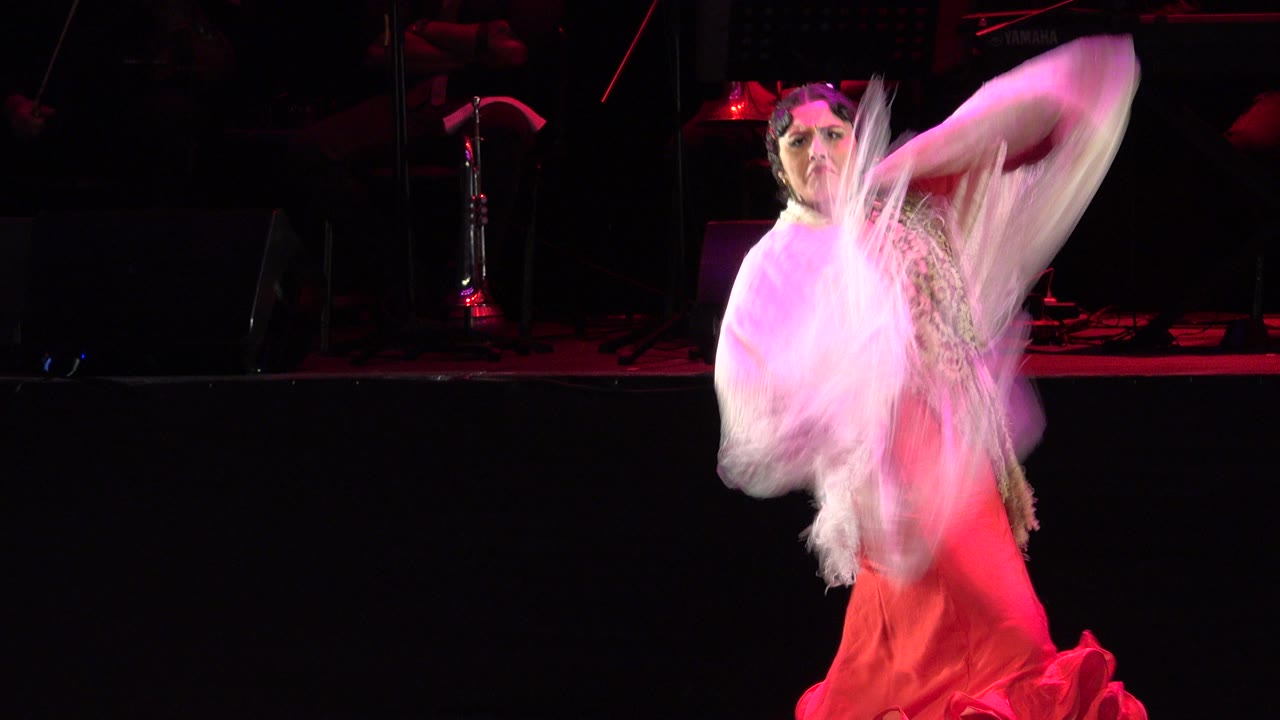 In Cabo San Lucas, Mexico, the dancer lifts the pale fringed shawl high across her upper body as her red gown stands out under pink stage light.