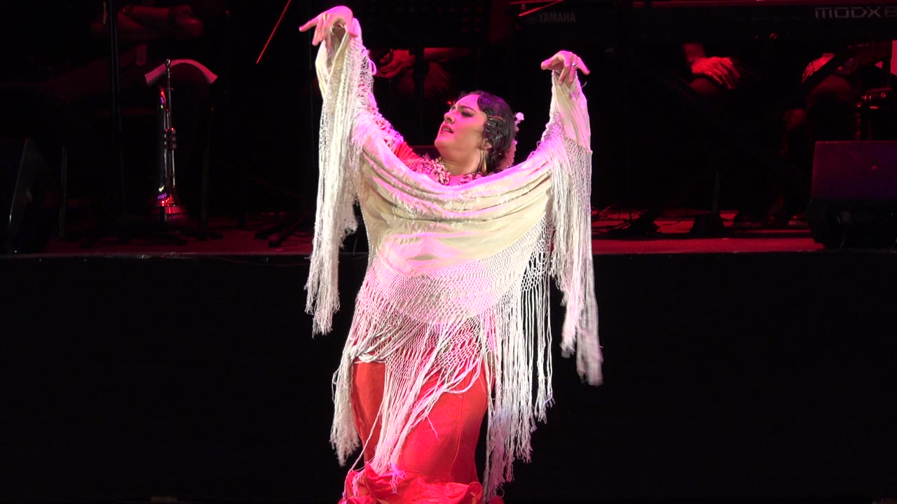 Adult woman in a red dress raises a white fringed shawl on a dark stage.