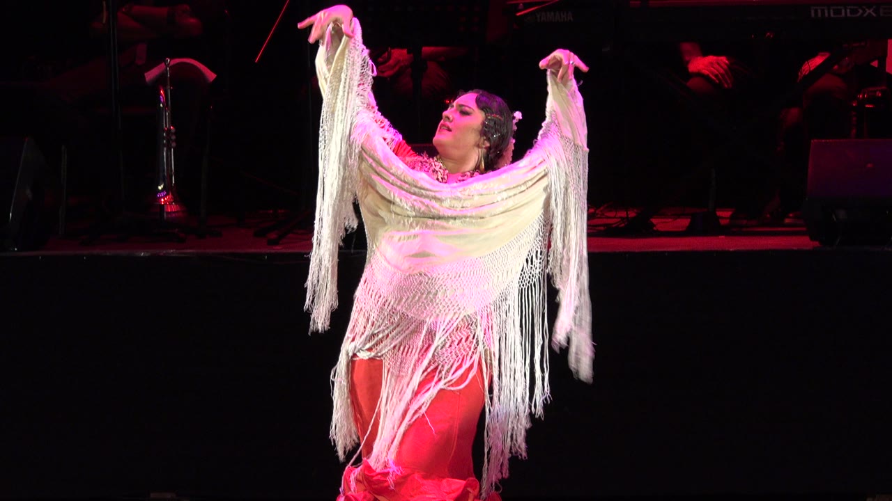 In Cabo San Lucas, Mexico, the adult dancer holds the white fringe high across her shoulders as red light catches the dress and stage floor.