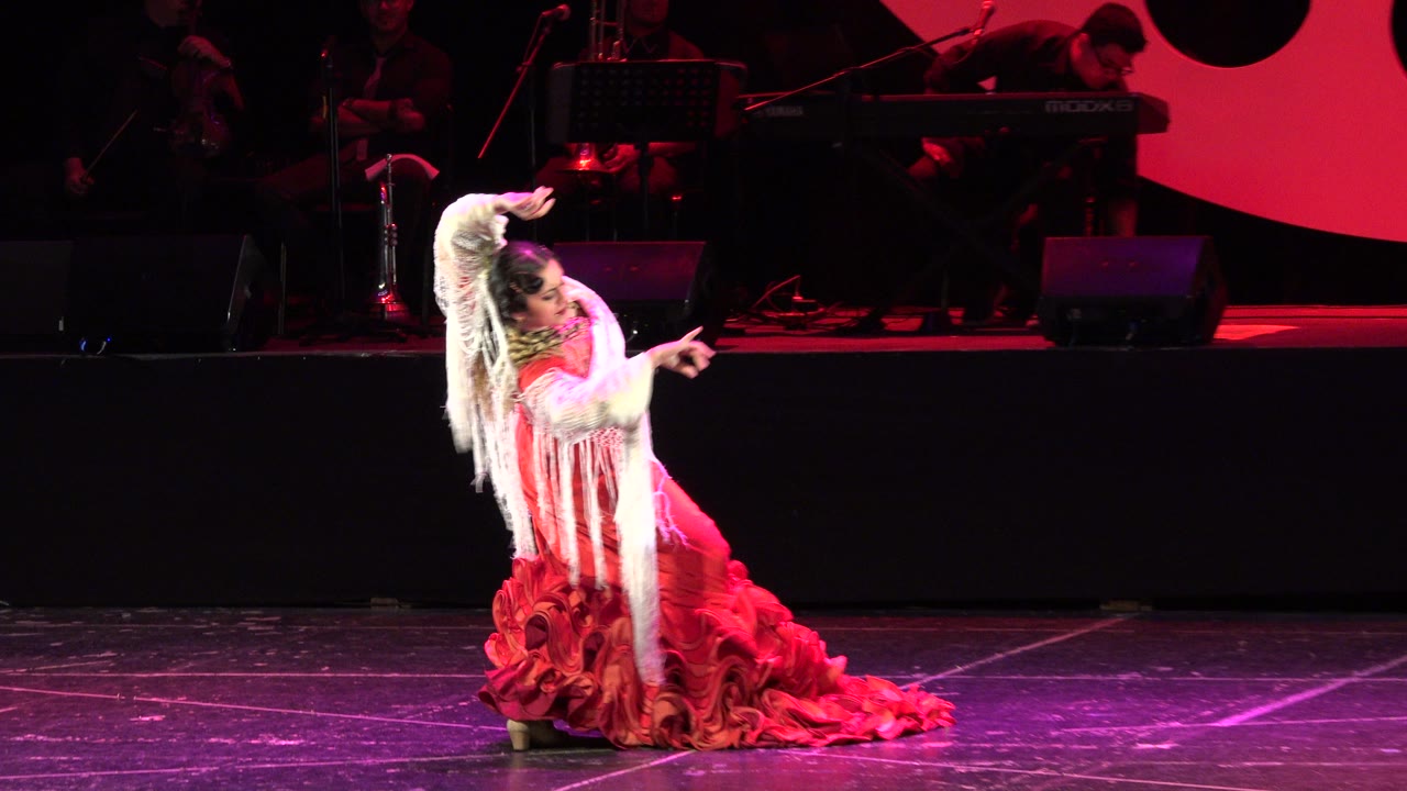 A female dancer in a red ruffled dress and white fringed shawl bends into a low pose on a red-lit stage in Cabo San Lucas, Mexico, with musicians dimly visible behind her.