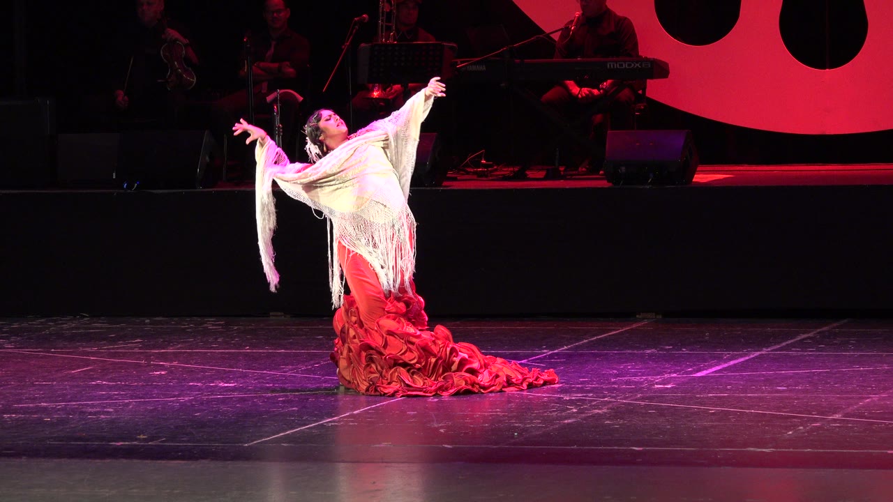 Woman in red dress and white shawl posing on a lit stage.